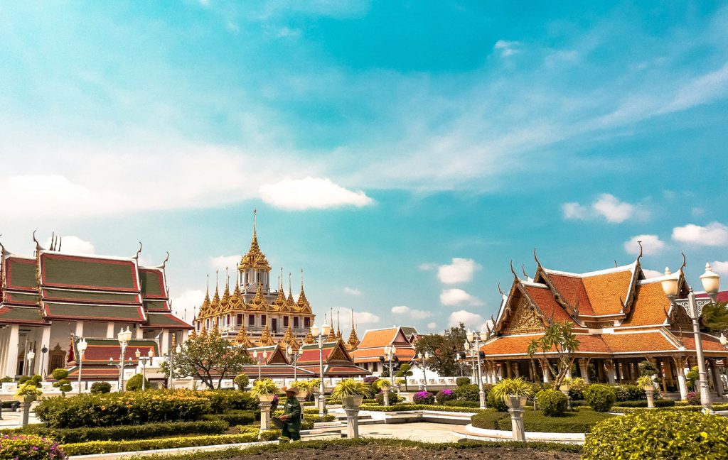 Ornate Thai temples with red roofs and golden spires stand under a bright blue sky.