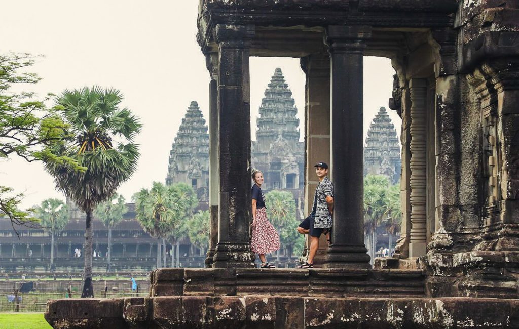 Two tourists stand among stone pillars with Angkor Wat’s iconic towers visible in the background.