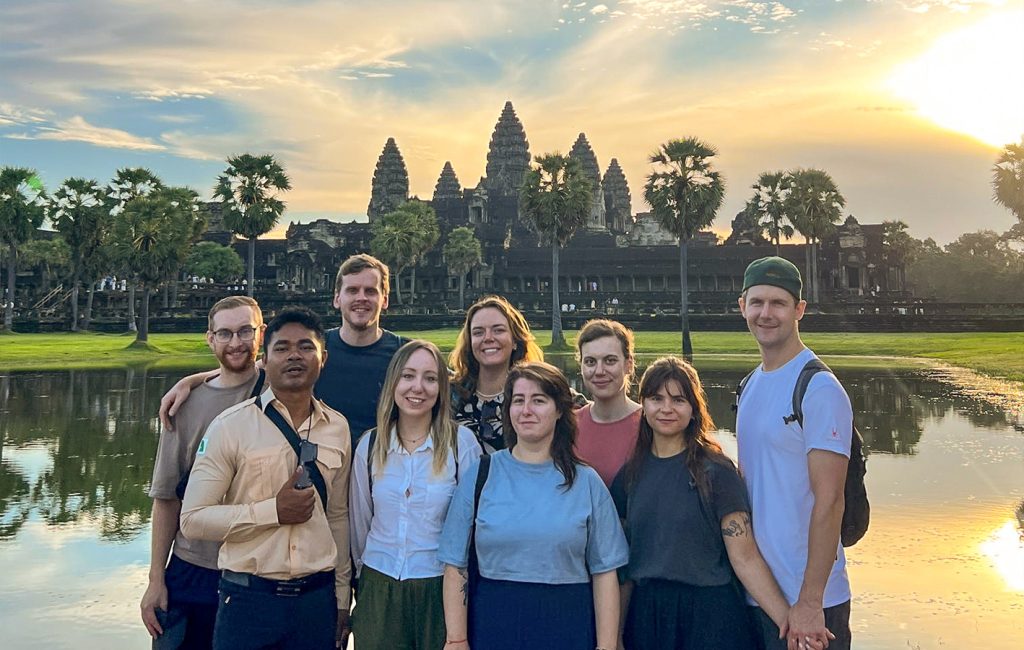 A group of smiling tourists pose in front of Angkor Wat at sunrise beside its reflective pond.