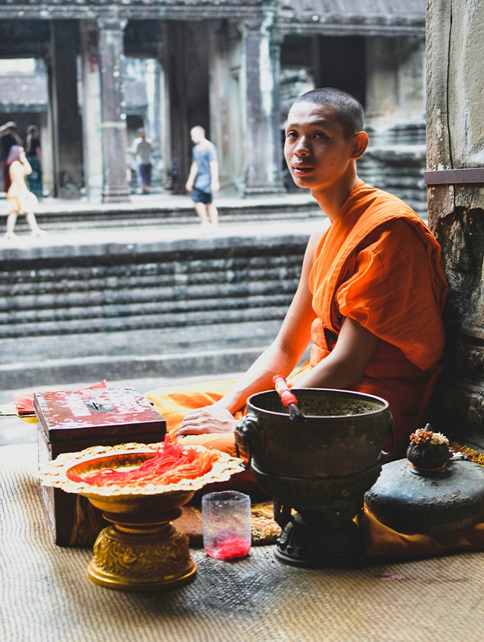 A young Buddhist monk in orange robes sits beside ceremonial items inside a temple corridor.