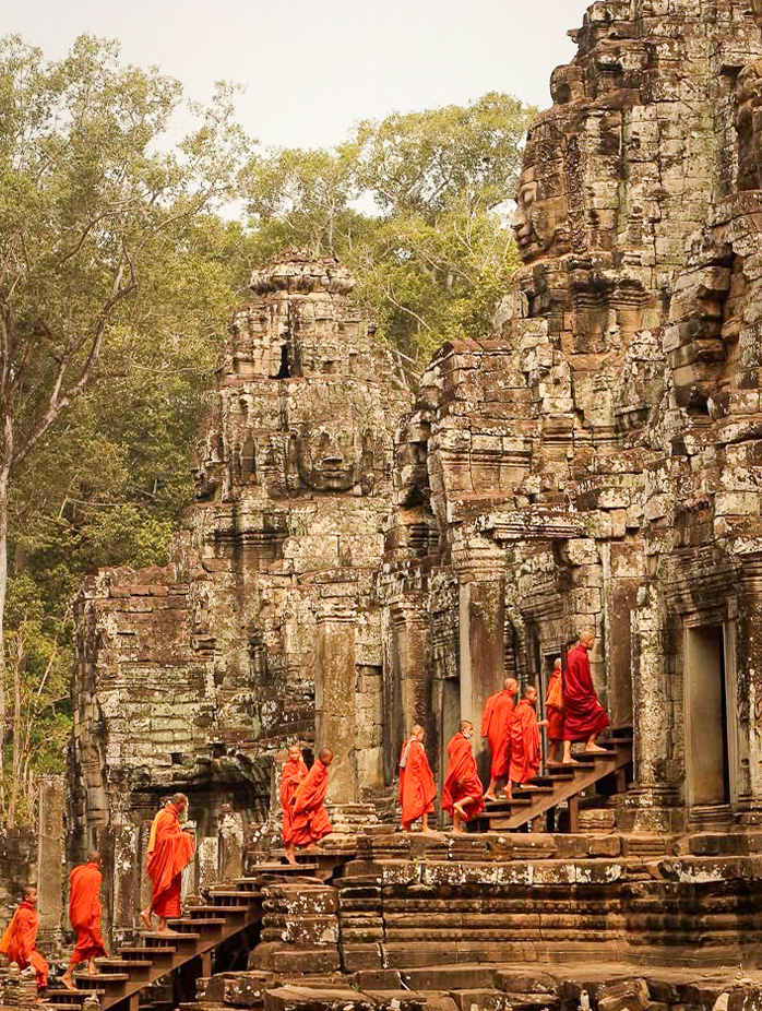 Buddhist monks in orange robes walk up ancient stone steps at Bayon Temple surrounded by carved faces.