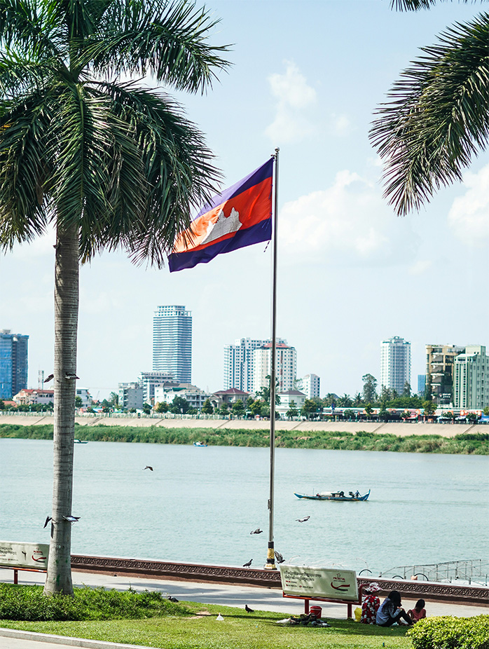 The Cambodian flag waves beside a riverside promenade with modern city buildings across the water.