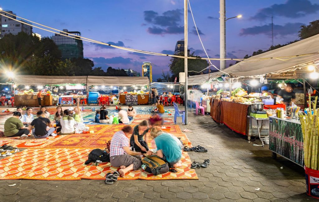People gather on patterned mats at an evening street market lined with food stalls and bright lights.