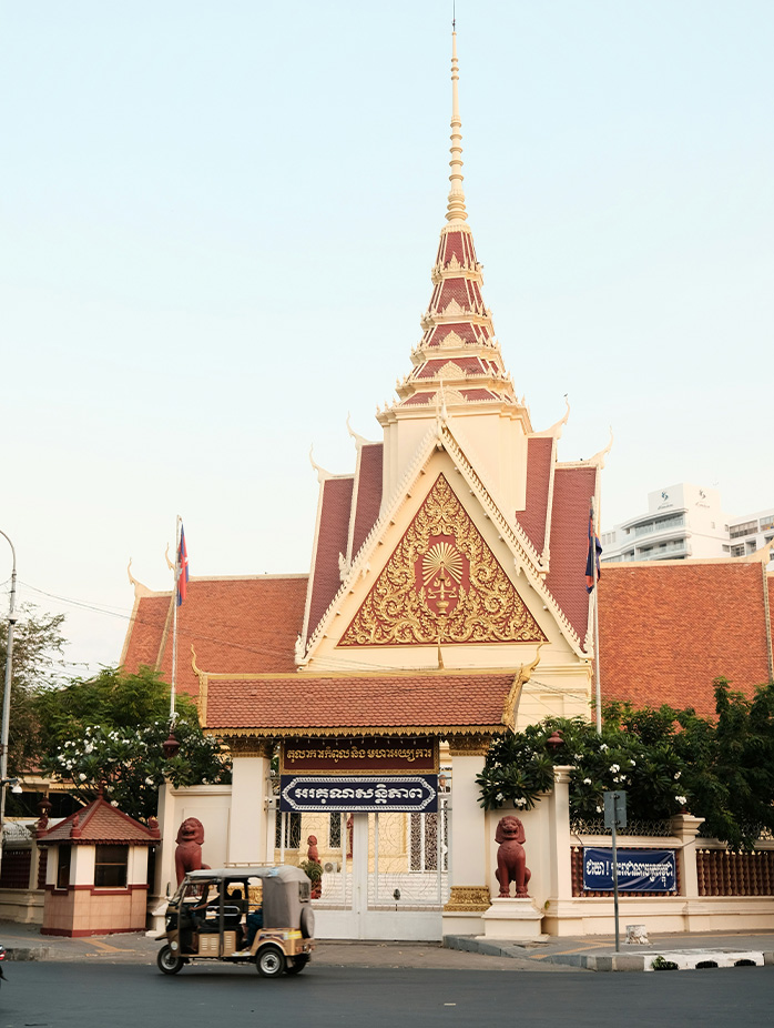 A traditional Cambodian temple with ornate red and gold details stands by a street where a tuk-tuk passes.