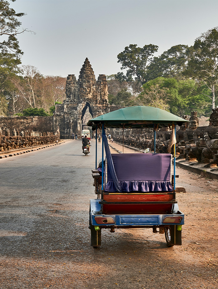 A tuk-tuk drives toward an ancient stone gate flanked by statues at Angkor Thom.