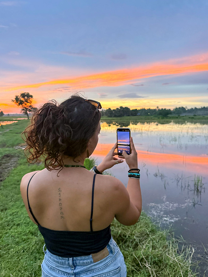 A woman photographs a colorful sunset reflected in a lakeside field.