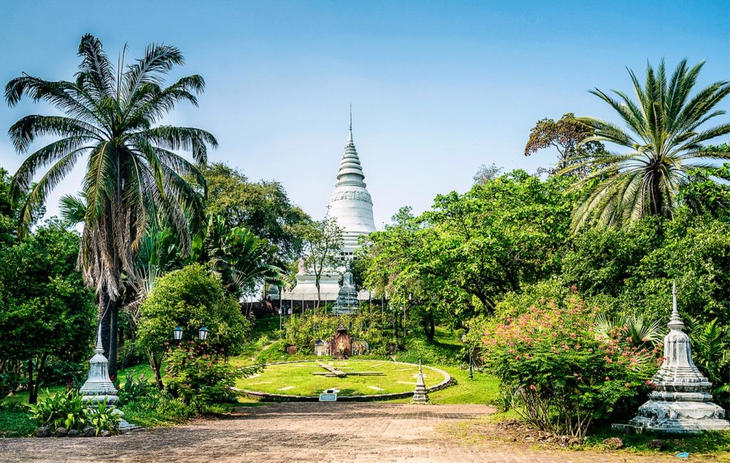 A white stupa rises above lush tropical gardens with palm trees under a bright blue sky.